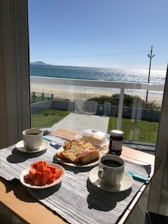 A dining table is set by a large window, offering a view of the sea. Two cups of coffee and a plate of sliced papaya accompany a slice of bread with jam. Natural light enhances the coastal scenery outside.