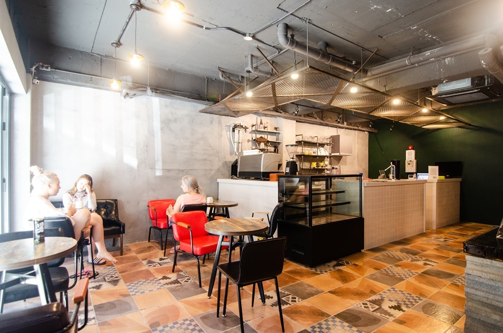 A modern communal space is depicted, featuring a variety of seating options including red armchairs and black chairs around small tables. An open kitchen area is visible in the background, with a counter displaying food and beverages. Soft lighting enhances the welcoming ambiance.