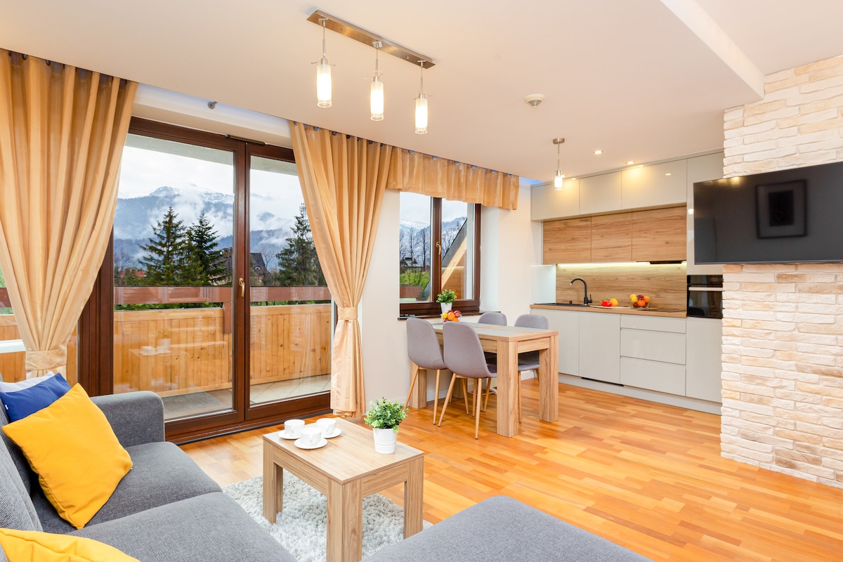 A modern living space features a gray sofa with decorative pillows facing a wooden table. Large windows provide mountain views and access to the balcony. The kitchenette and dining area, equipped with a table and chairs, are illuminated by recessed lighting.
