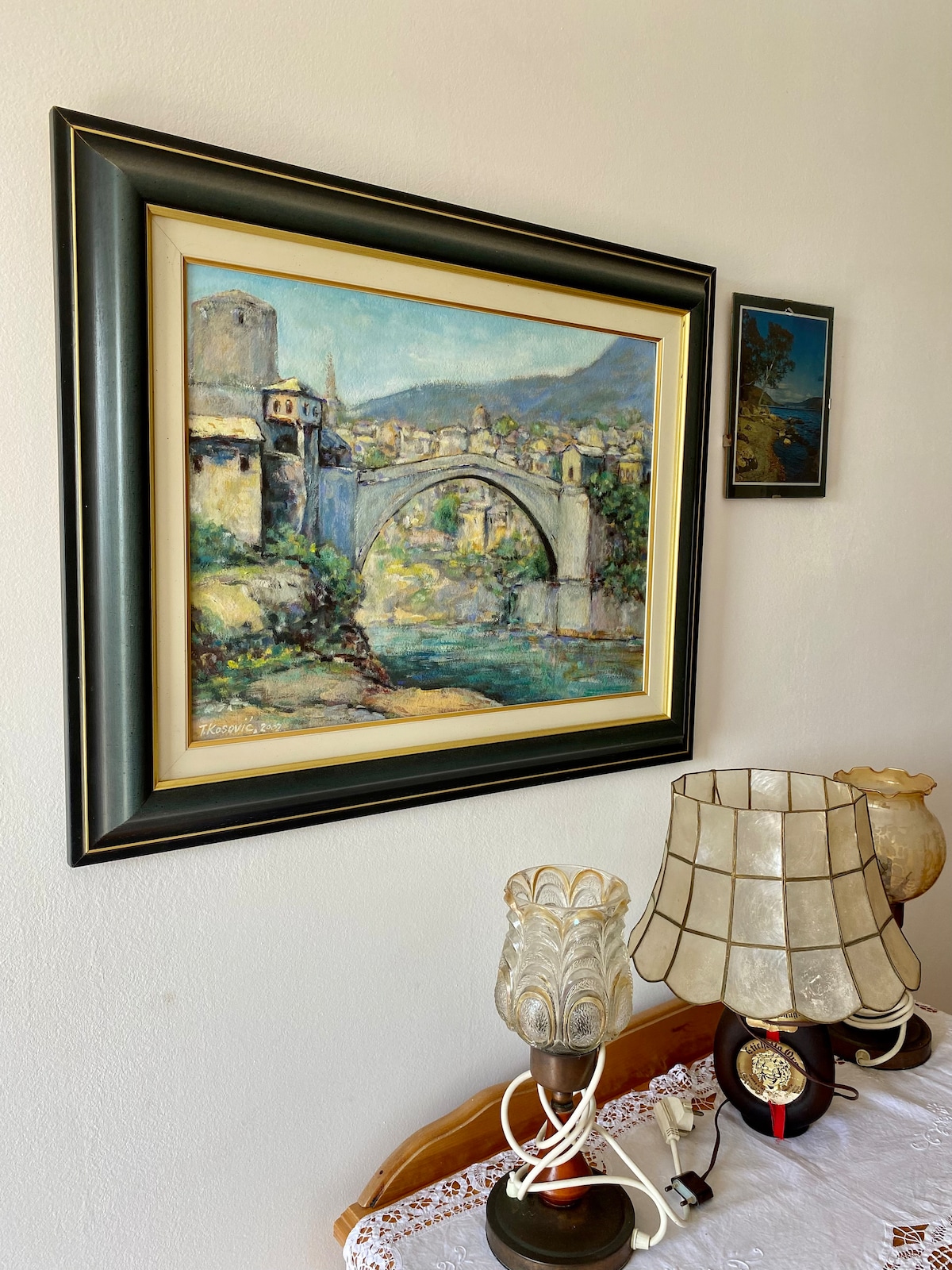 A framed painting of a bridge over a river is displayed on a light-colored wall. A small framed artwork is visible beside it. In the foreground, a wooden table is adorned with a white lace tablecloth, featuring two decorative lamps with textured glass shades.