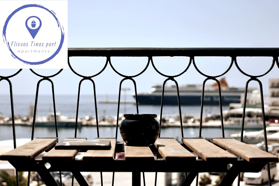 A wooden balcony table is positioned with an unobstructed view of the bustling port, featuring docked boats and a large ship in the background. The scene reflects a serene atmosphere ideal for enjoying drinks or meals outdoors.
