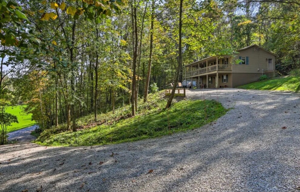 A gravel driveway winds through greenery, leading to a two-story home nestled among trees. The house features covered porches on both levels, providing inviting outdoor spaces. Sunlight filters through the foliage, highlighting the natural surroundings.