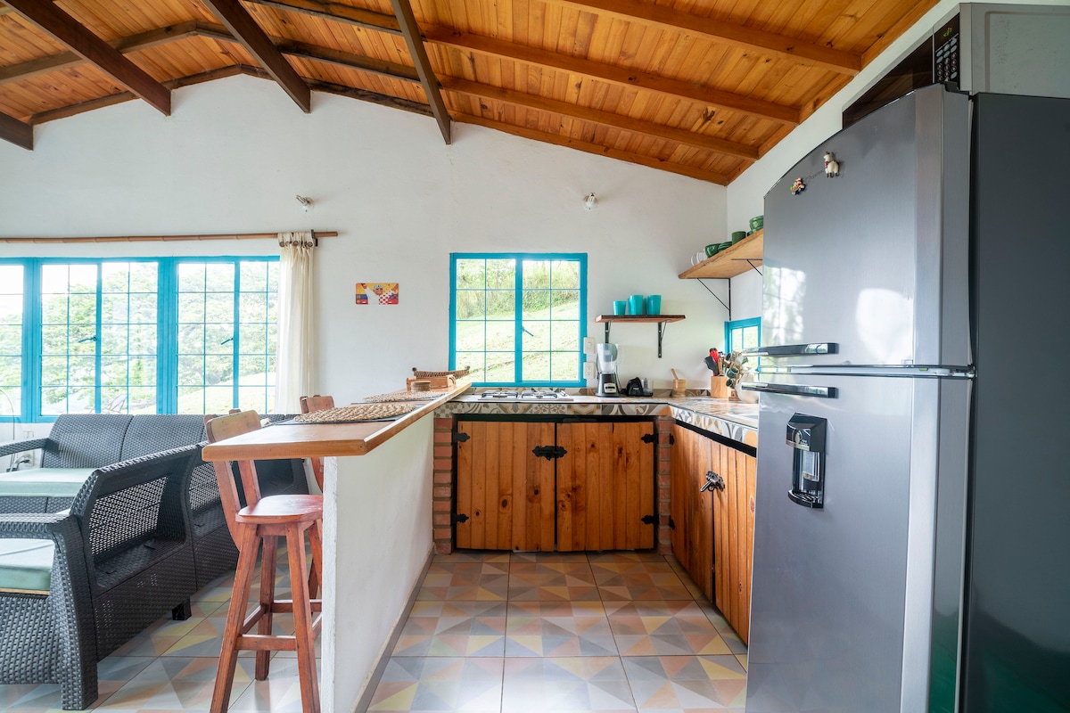 The kitchen features wooden cabinetry and a granite countertop. Large windows allow natural light to fill the space, highlighting the tiled floor. A sturdy refrigerator stands tall, while a wooden bar area offers additional seating. The cozy dining area is visible beyond the kitchen.