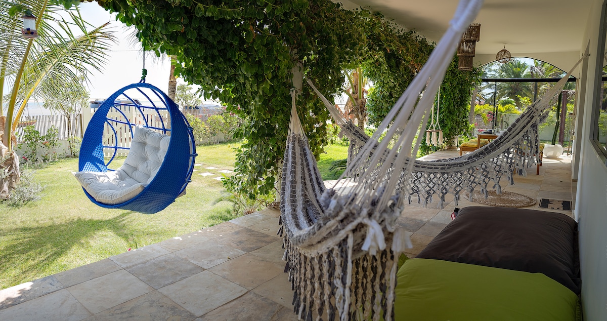 A spacious terrace showcases several hammocks and a blue hanging chair, surrounded by lush greenery and vibrant garden views. Comfortable cushions provide a relaxing setting, while natural light filters through the trees, creating a serene ambiance perfect for unwinding.