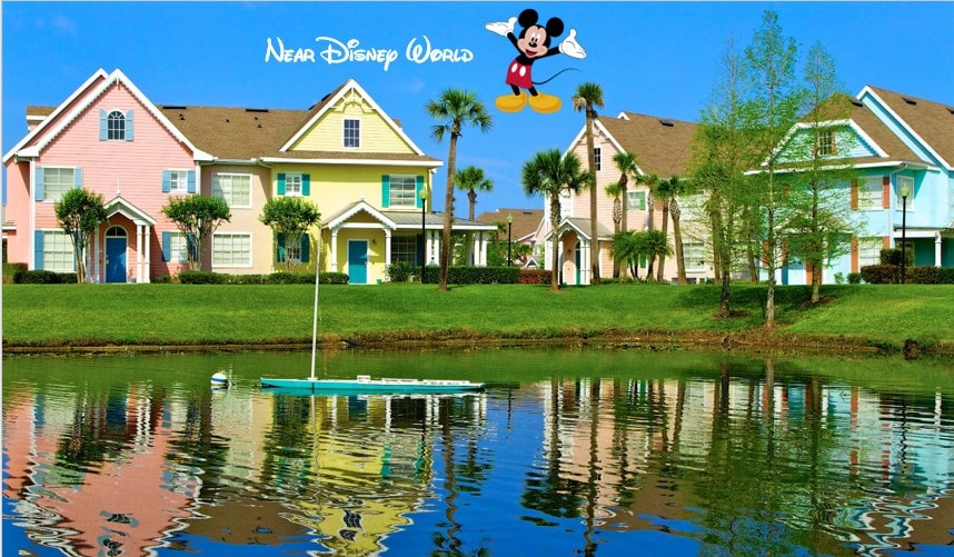 A serene reflection of colorful townhouses is visible on the calm water of the pond in the foreground. Palm trees line the shore, adding a tropical touch to the scenic landscape under a clear blue sky.