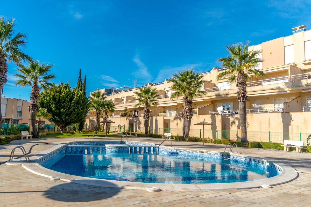 A large communal pool is surrounded by a paved area and lush palm trees, creating an inviting outdoor space. The sunlit sky reflects off the water, and nearby structures offer additional seating options. The setting evokes a relaxed atmosphere for residents to enjoy.