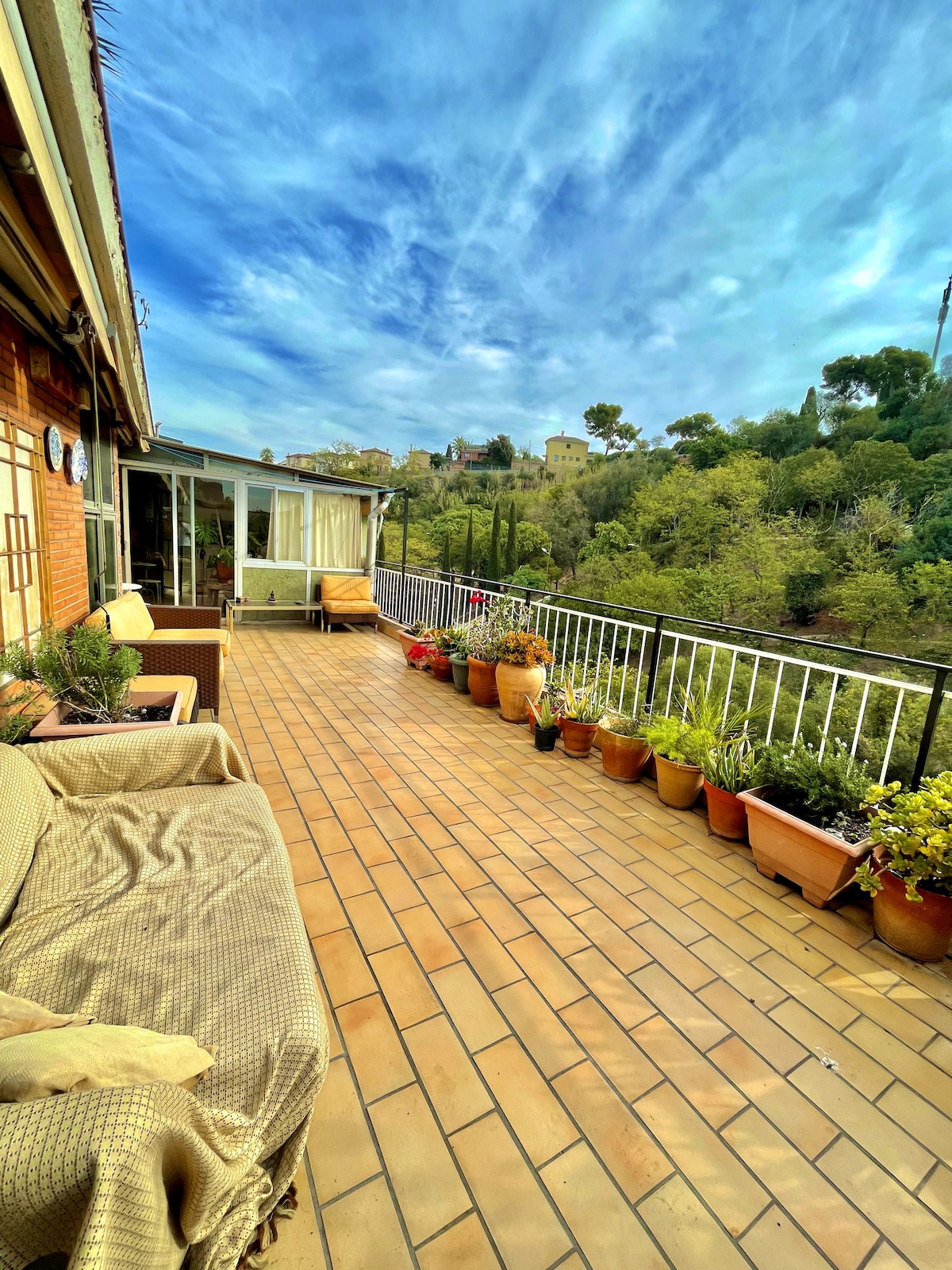 A spacious terrace is adorned with potted plants and flowers, showcasing vibrant greenery against a natural backdrop. Comfortable outdoor seating is provided, inviting relaxation. The sky above is filled with soft clouds, enhancing the serene atmosphere of this outdoor space.