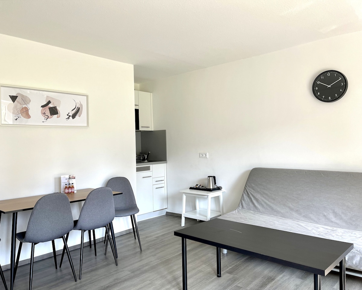 The living area features a simple gray sofa bed with a smooth surface and a black coffee table. A set of three dining chairs is positioned around a small table. The kitchenette is seen in the background, providing essential appliances, and a wall clock is mounted above the sofa.