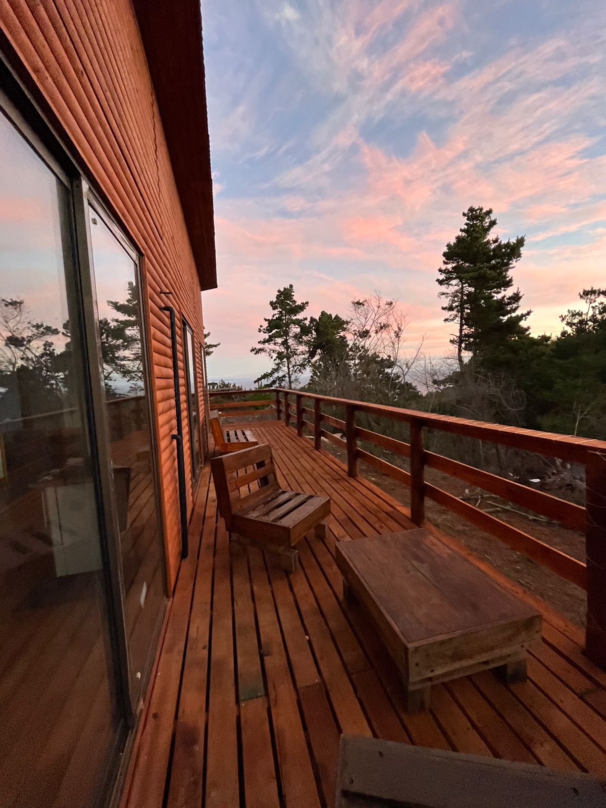 A spacious wooden deck is complemented by several outdoor seating options, including two lounge chairs and a coffee table. The warm tones of the wood contrast with the colorful sunset sky, while trees frame the serene view of the surrounding landscape.