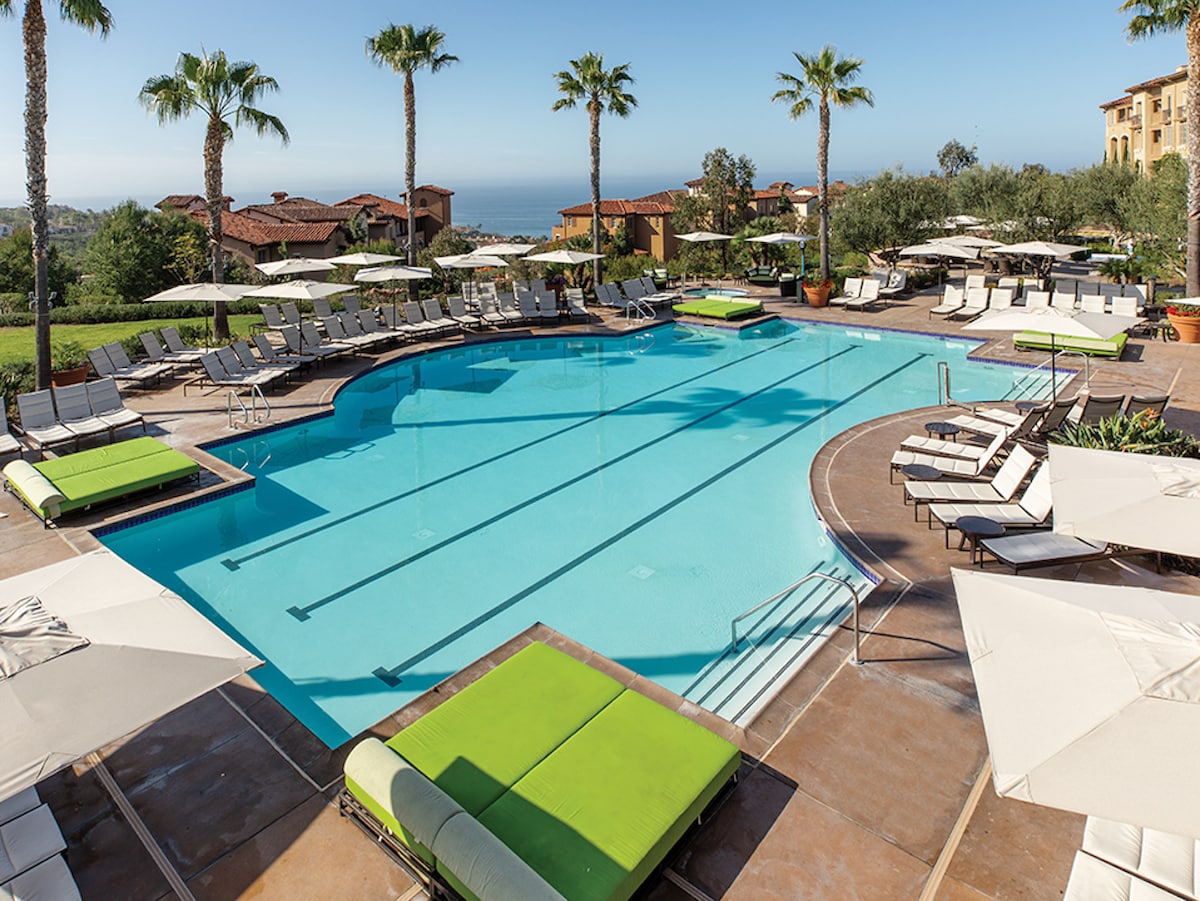 An outdoor pool area is surrounded by lounge chairs and umbrellas. Palm trees provide shade beside the turquoise pool, where bright green cushions add vibrant color. The backdrop features terracotta-tiled buildings, complementing the lush landscape and expansive ocean views.