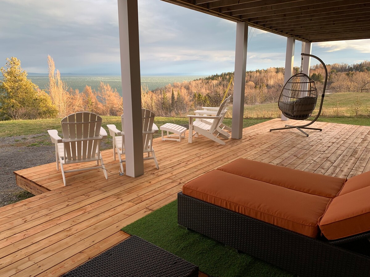 A spacious wooden deck features a cozy seating arrangement, including a hanging chair and two loungers. A large cushioned lounge area is complemented by lush green grass. The panoramic view of the landscape extends towards the horizon, framed by autumn foliage.
