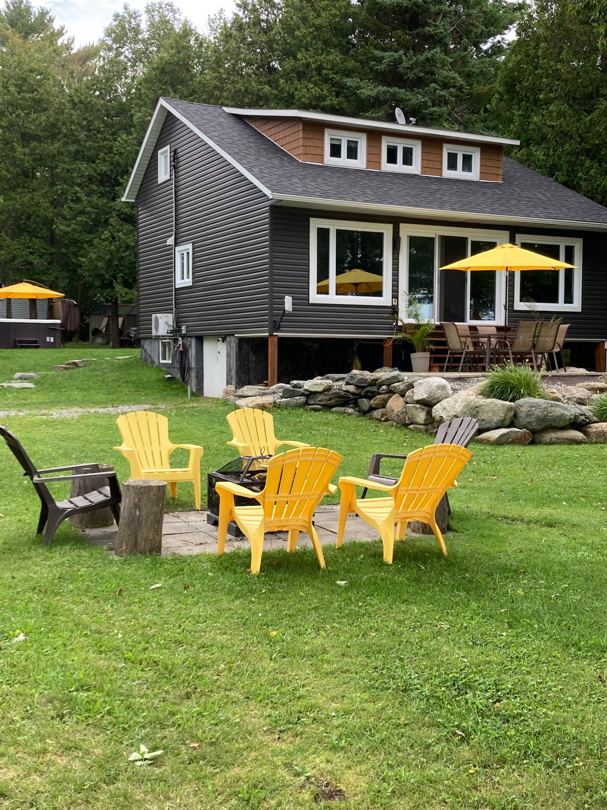 The exterior of the chalet is shown, featuring yellow Adirondack chairs arranged around a fire pit on a grassy area. Large windows provide views of the interior, while a stone pathway leads to a patio with dining furniture and umbrellas for shade.