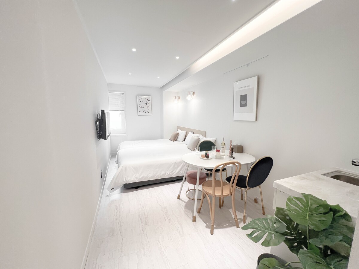 A modern studio space features a spacious bed with crisp linens, accompanied by a small dining table set for two. Natural light filters through a nearby window, highlighting the minimalist décor and soft grey walls. A small kitchenette is neatly integrated into the room.