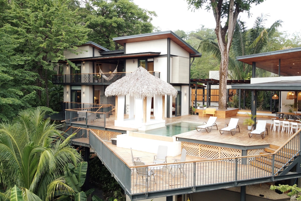 The villa features a raised wooden deck surrounded by lush greenery, with a swimming pool at its center. A traditional thatched-roof 'Rancho' is visible, along with various lounge chairs arranged for relaxation. The two-story structure integrates seamlessly with nature, providing an inviting atmosphere.