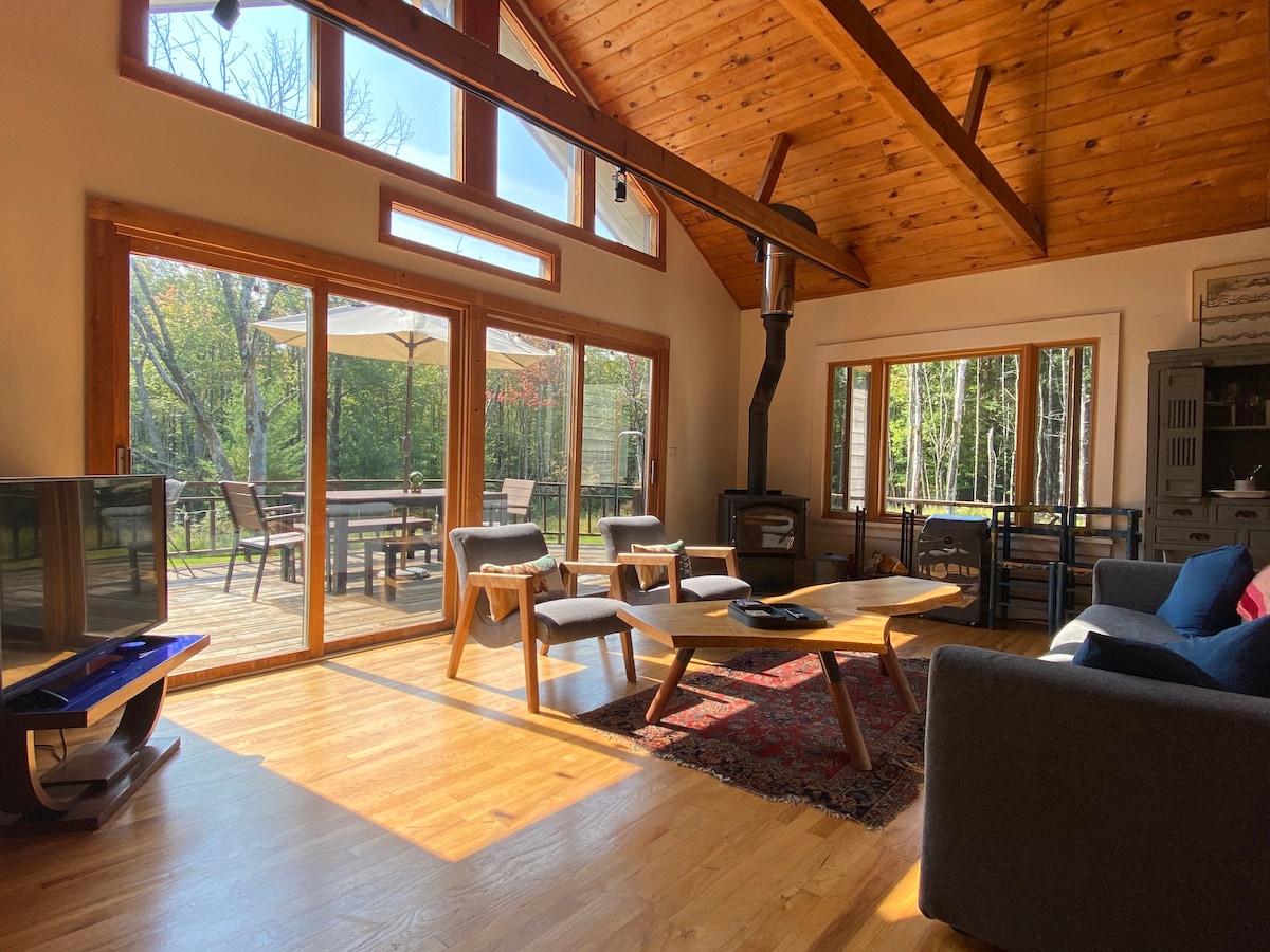 An open living space features vaulted wooden ceilings and large sliding glass doors that provide natural light. A cozy seating area includes a sofa and wooden coffee table. A wood-burning stove is positioned nearby, surrounded by warm wooden elements and a view of the deck outside.