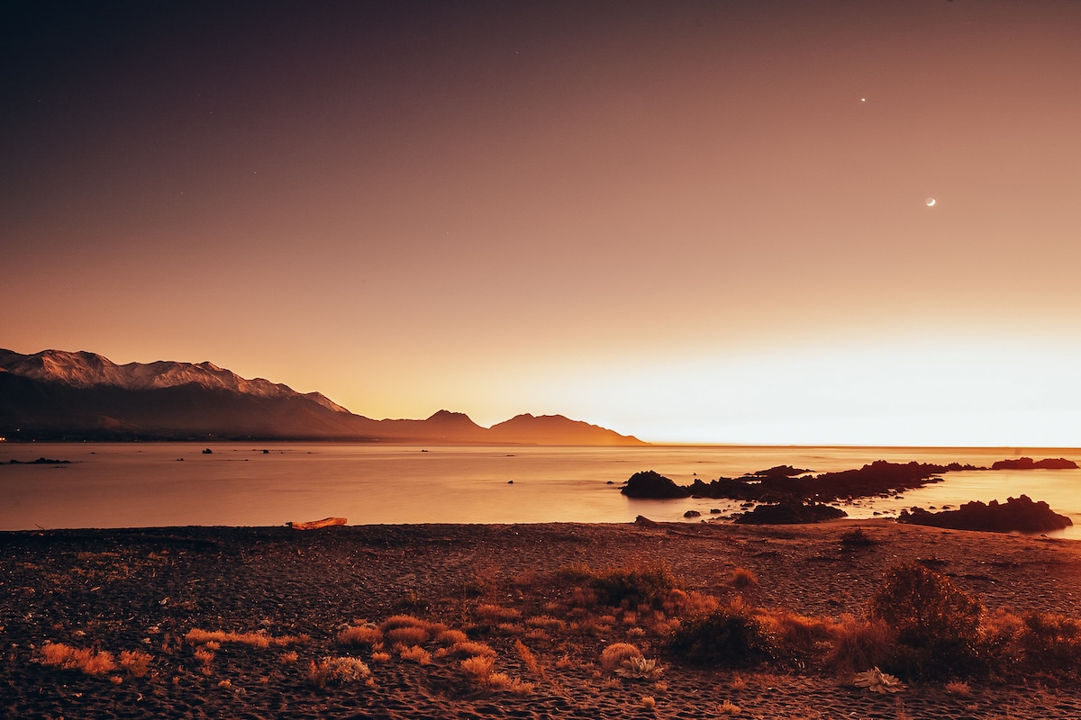 A serene beachfront scene captures a muted sunset over calm waters, with gentle waves lapping at the shore. Silhouettes of distant mountains create a dramatic backdrop against the soft gradient of orange and purple hues in the sky.