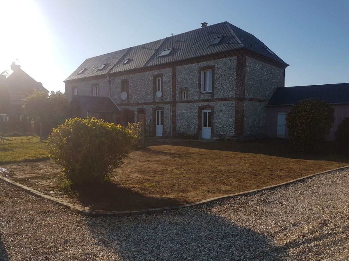 A rustic stone house features a symmetrical design with multiple windows and a steep roof. A grassy area surrounds the building, bordered by neatly laid gravel pathways. Sunlight bathes the exterior, highlighting the texture of the stonework and the manicured shrubbery.