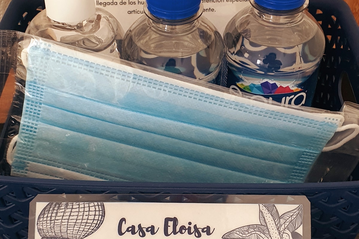 A basket containing essential items for guests is shown. Included are three bottles of purified water, a hand sanitizer, a disposable face mask, and a welcome note indicating the name 'Casa Eloisa.'