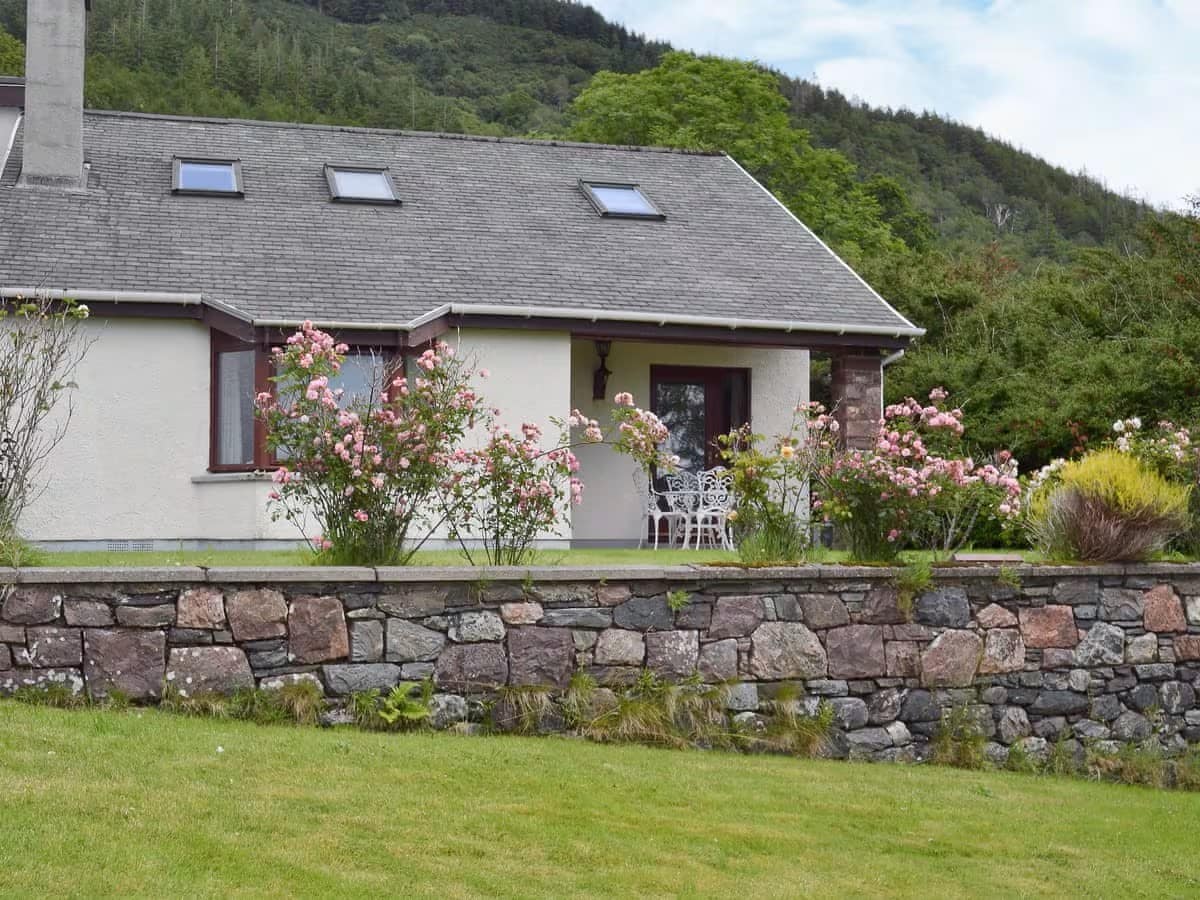 A charming cottage is set against a backdrop of lush greenery and hills. The exterior features a stone wall with blooming rose bushes, white chairs, and a small table on a patio. Skylights allow natural light to illuminate the roof.