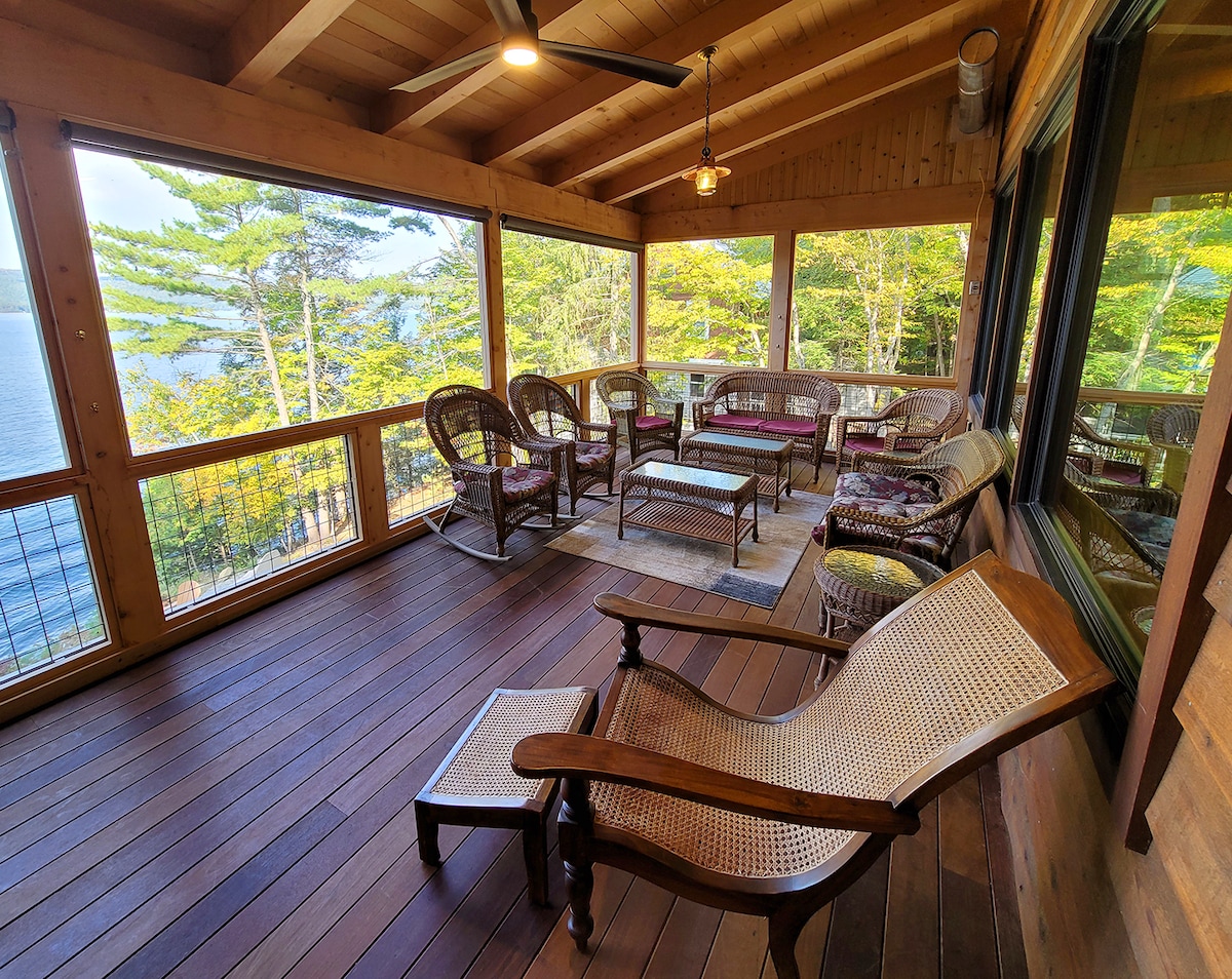A tranquil screened porch features wooden furnishings, including several woven chairs and glass-topped tables. Large windows offer a view of the surrounding trees and the lake, with natural light filtering in, creating a relaxing space to enjoy the outdoors.