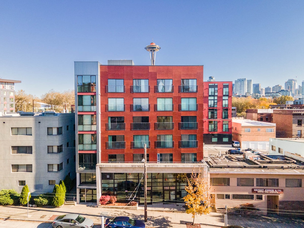 The exterior view of a modern building is captured, showcasing a blend of red and grey façades. Large windows reflect the sky and surrounding city, with the iconic Space Needle visible in the background. The building's entrance features a welcoming awning and landscaped greenery.