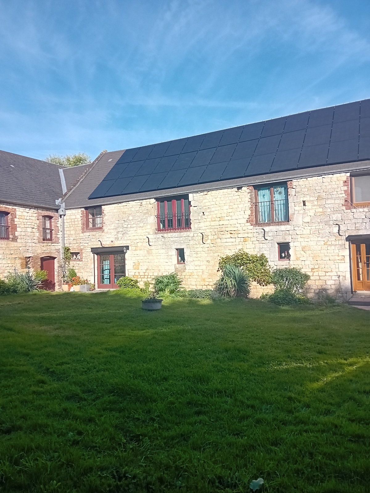 A stone building features a sloped roof adorned with solar panels and vibrant windows framed by natural greenery. A well-maintained grassy area surrounds the structure, enhancing the rural setting. The architecture reflects a blend of rustic charm and modern sustainability.