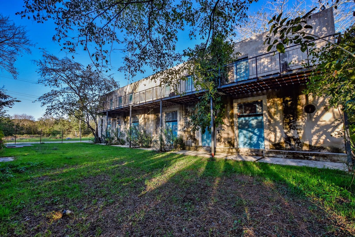 An exterior view of a two-story loft in a historic hacienda, surrounded by lush greenery. Balconies with blue doors are visible, offering a glimpse into outdoor living. Large trees provide shade over the well-kept grass, creating a peaceful atmosphere.