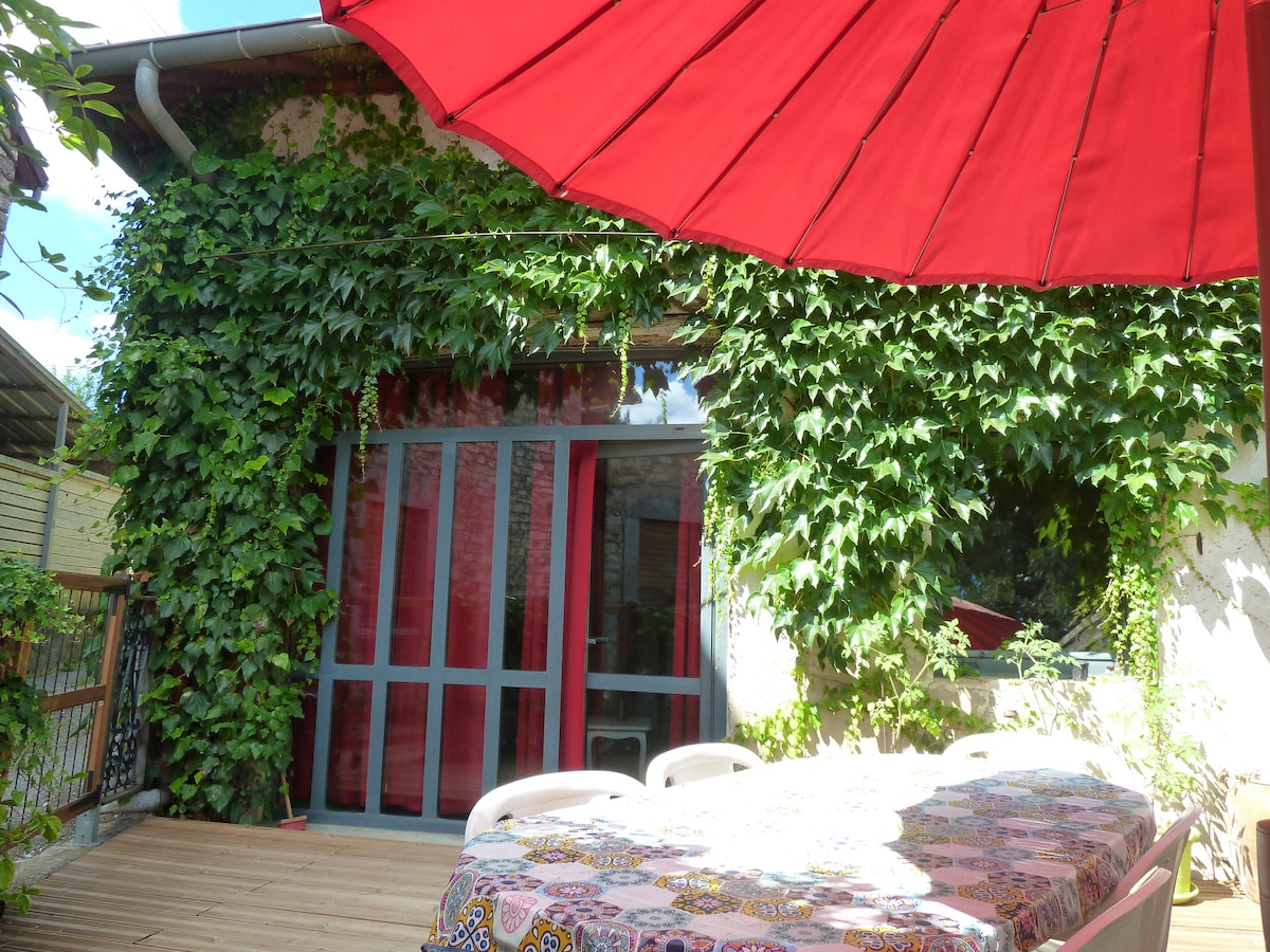 A wooden terrace is covered with a vibrant patterned tablecloth, surrounded by lush green ivy. A large red umbrella provides shade, while glass doors in the background offer access to the interior space.