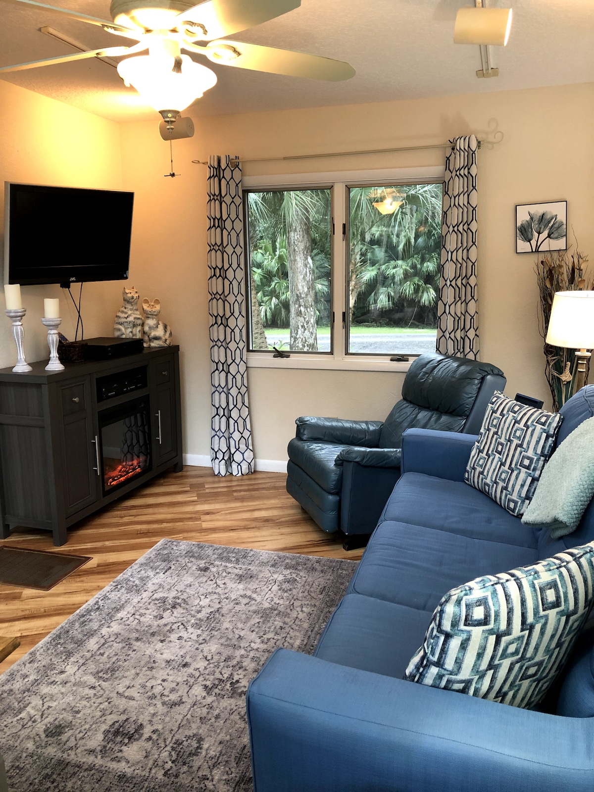 A living area is furnished with a teal seating arrangement, including a cozy couch and a recliner. A mounted television is positioned above a media console. Natural light enters through a window adorned with patterned curtains, while a decorative rug complements the wooden flooring.