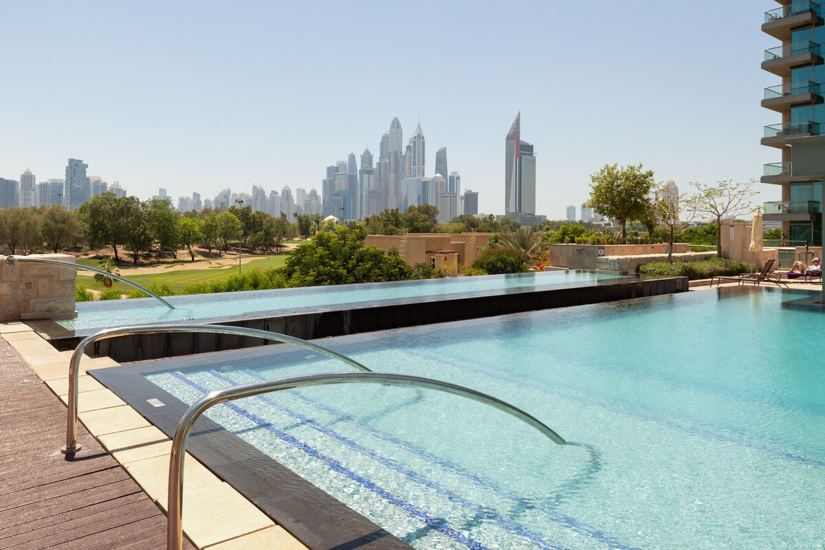 A sparkling infinity pool extends towards a skyline of tall buildings, framed by lush greenery and a golf course. Sunlight reflects off the water's surface, creating a tranquil scene that invites relaxation.
