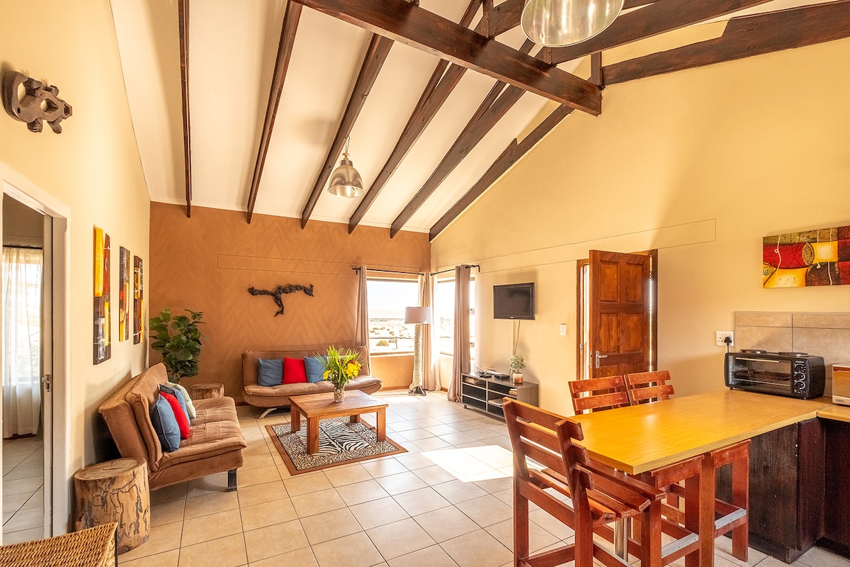 A spacious living area features high ceilings with exposed beams. Comfortable seating is arranged around a central coffee table. A dining area is highlighted by a wooden table and chairs. Large windows provide natural light, creating an inviting atmosphere.