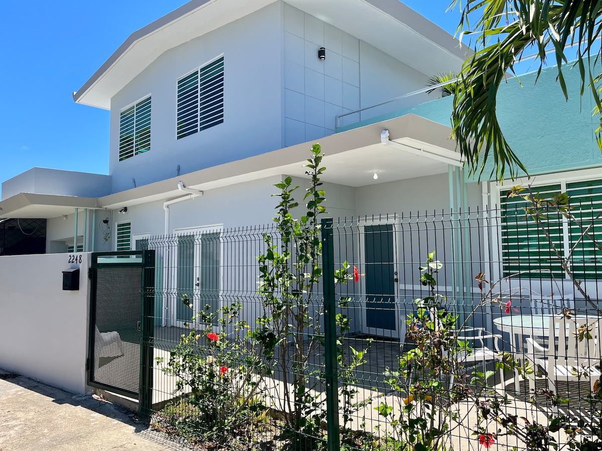 The exterior of a two-story beach house is presented, featuring a light-colored facade with green accents. A gated entrance and a patio area with outdoor seating are visible. Lush greenery and colorful flowers line the pathway, enhancing the welcoming atmosphere.
