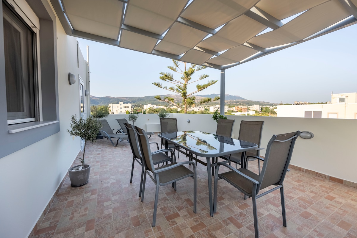 A spacious terrace features a large glass table surrounded by six chairs. A sunshade elegantly covers the seating area. Potted plants are positioned along the edge, with a partial view of distant mountains visible in the background.