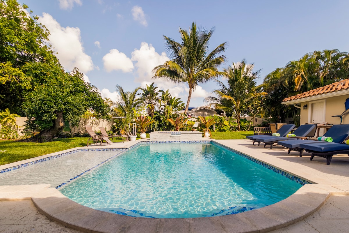 A serene backyard oasis features a spacious pool surrounded by a stone deck. Comfortable lounge chairs are positioned on one side, while lush tropical plants and palm trees create a tranquil environment. Sunlight reflects off the clear water, inviting relaxation and outdoor enjoyment.