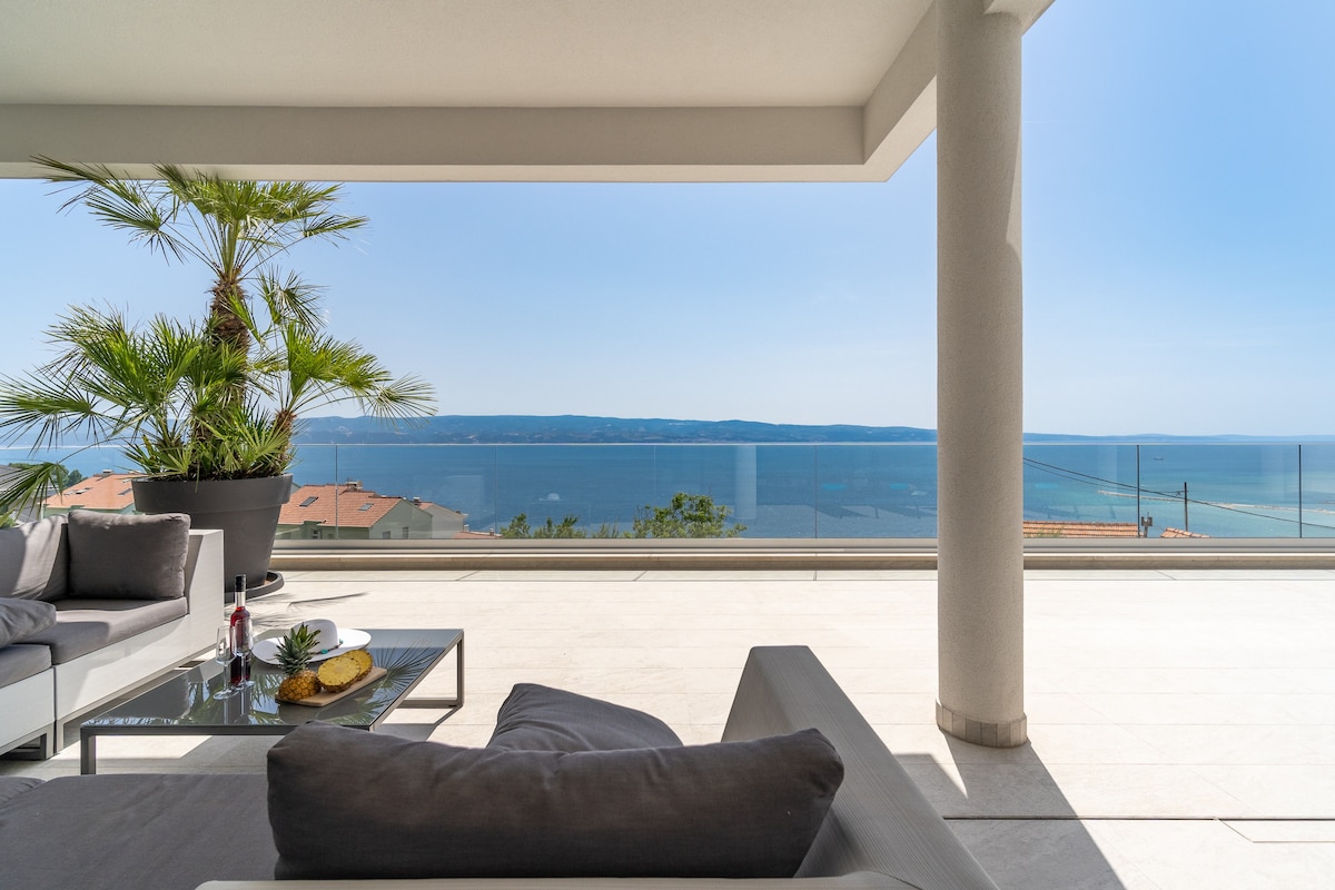 An outdoor terrace is visible, featuring lounge furniture arranged around a coffee table. A tall plant adds a touch of greenery. Panoramic sea views are framed by glass railings, with clear blue skies enhancing the serene atmosphere.