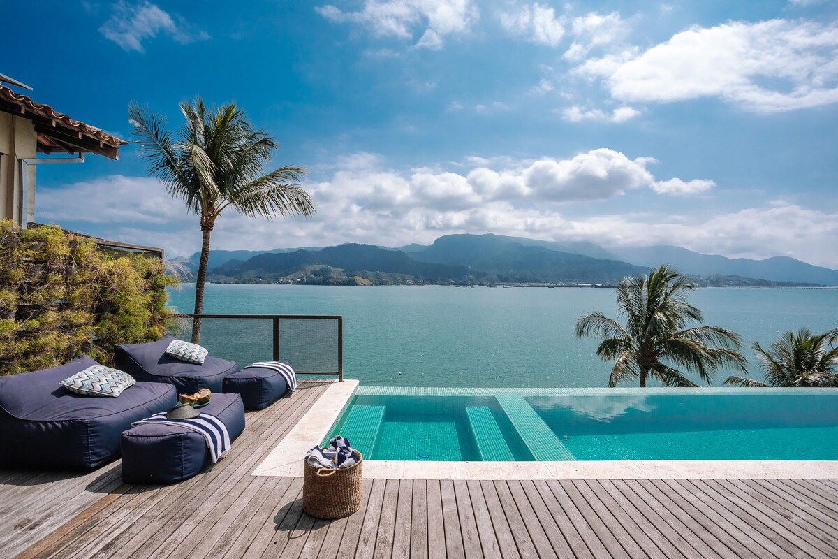 A sunlit pool area overlooks a serene body of water, featuring an infinite edge that creates a seamless view of the horizon. Comfortable lounge chairs are positioned nearby, with palm trees swaying gently in the breeze, enhancing the tranquil coastal ambiance.
