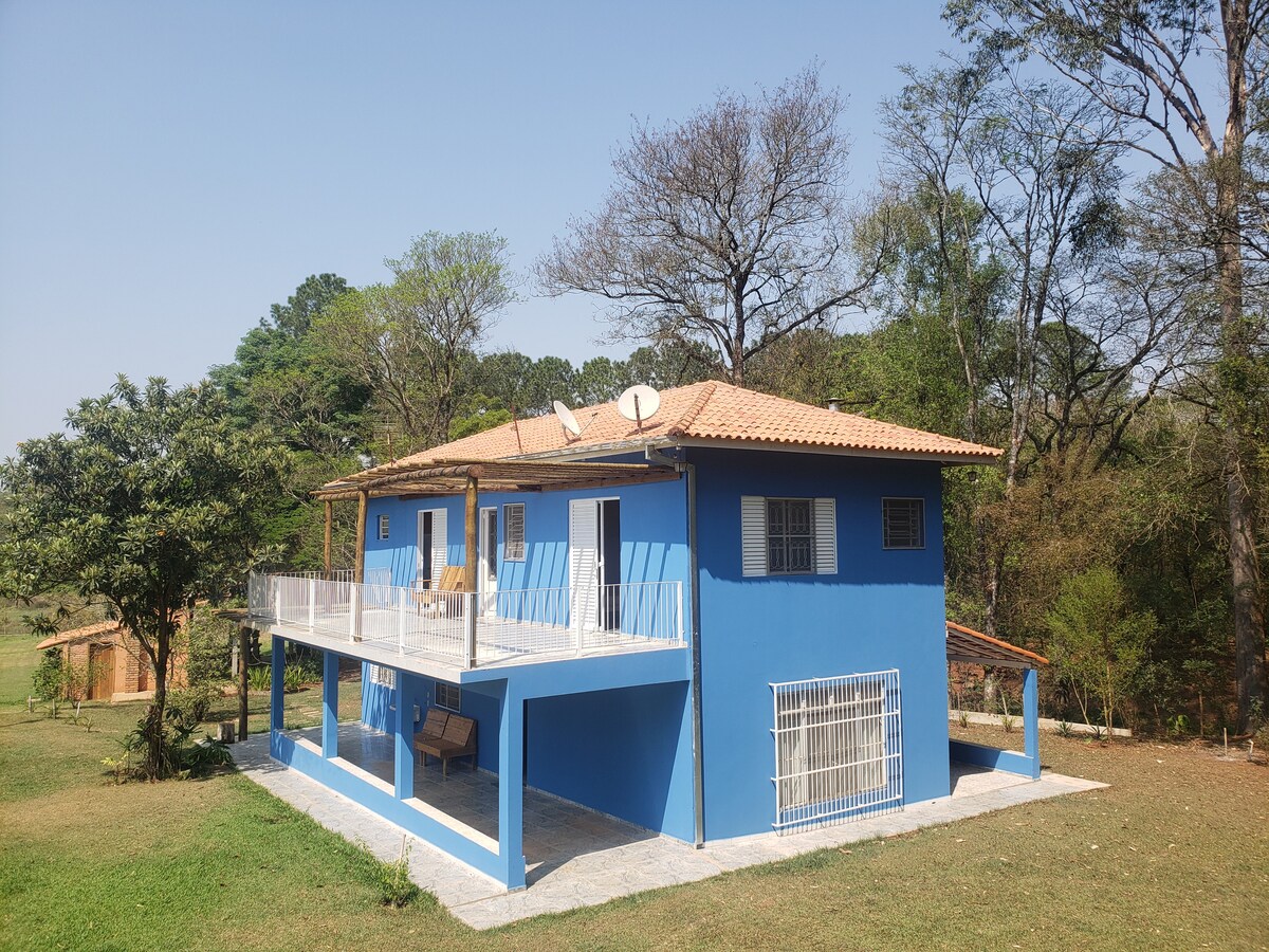 A two-story blue house is set against a backdrop of trees and clear sky. A spacious porch extends from the front, featuring seating arrangements. Satellite dishes are visible on the roof, complementing the rural landscape that surrounds the property.