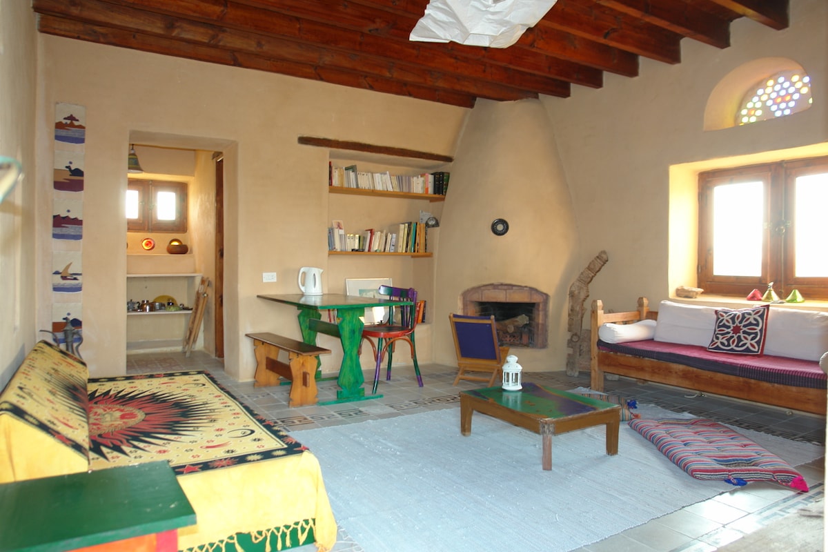 The living room features a rustic design with wooden beams overhead and a cozy seating area. A circular table and colorful cushions invite relaxation. A small bookshelf is present, alongside a large window adorned with stained glass, allowing natural light to fill the space.