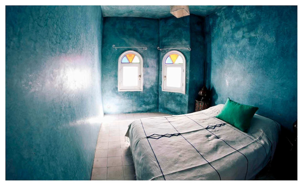 A tranquil bedroom features vibrant blue walls and two arched windows adorned with colored panes. A spacious bed is positioned against the wall, with a green accent pillow adding a pop of color. Natural light is softly diffused through the windows, creating a serene atmosphere.