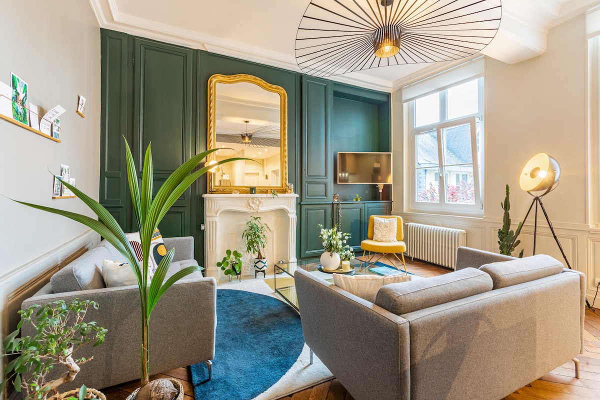 A stylish living area features a large mirror above a decorative fireplace. Two gray sofas are positioned around a circular rug, accompanied by modern decor elements including a floor lamp and potted plants. Natural light fills the space through large windows, accentuating the green accent wall.