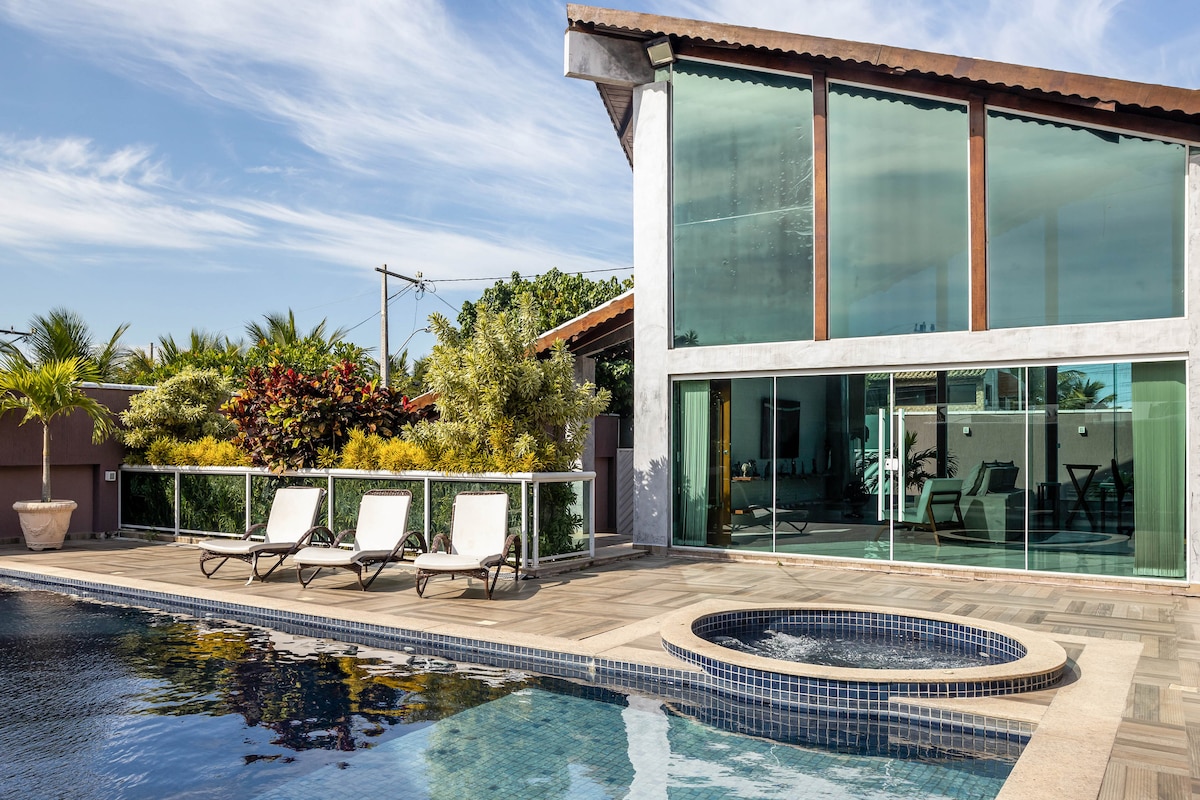 A modern house features large glass windows that provide views of the pool area. Lounge chairs are positioned around the pool, alongside an adjacent hot tub. Lush greenery is visible in the background, enhancing the outdoor space.