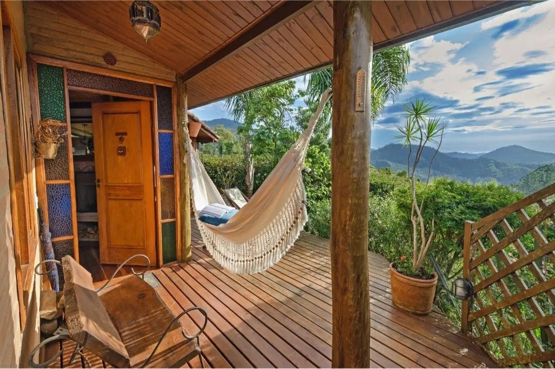 An inviting outdoor deck is furnished with a hammock and a small wooden table. Lush greenery and mountain views create a serene backdrop. The space is framed by wooden beams and features colorful glass details on the walls, enhancing its natural charm.
