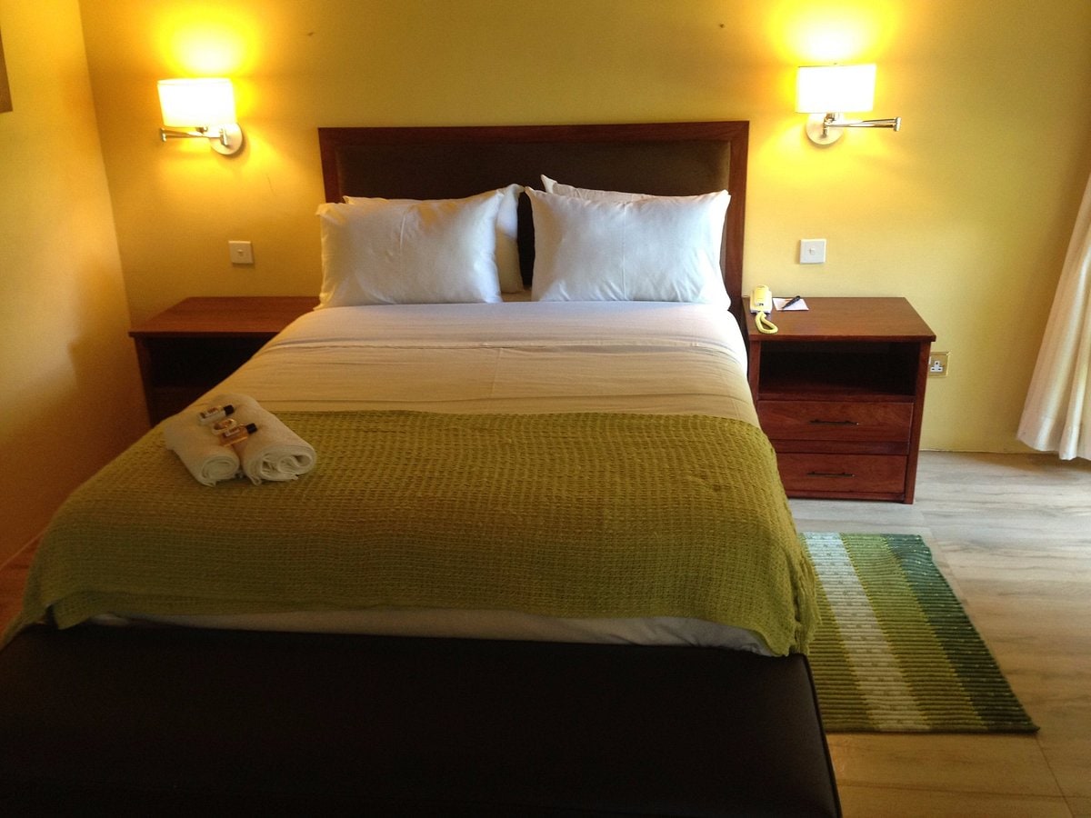 A cozy sleeping area features a spacious bed with crisp white linens and plush pillows. A bright green blanket is casually draped over the bed. Two bedside tables with lamps are positioned on either side, and a green accent rug is placed on the floor.