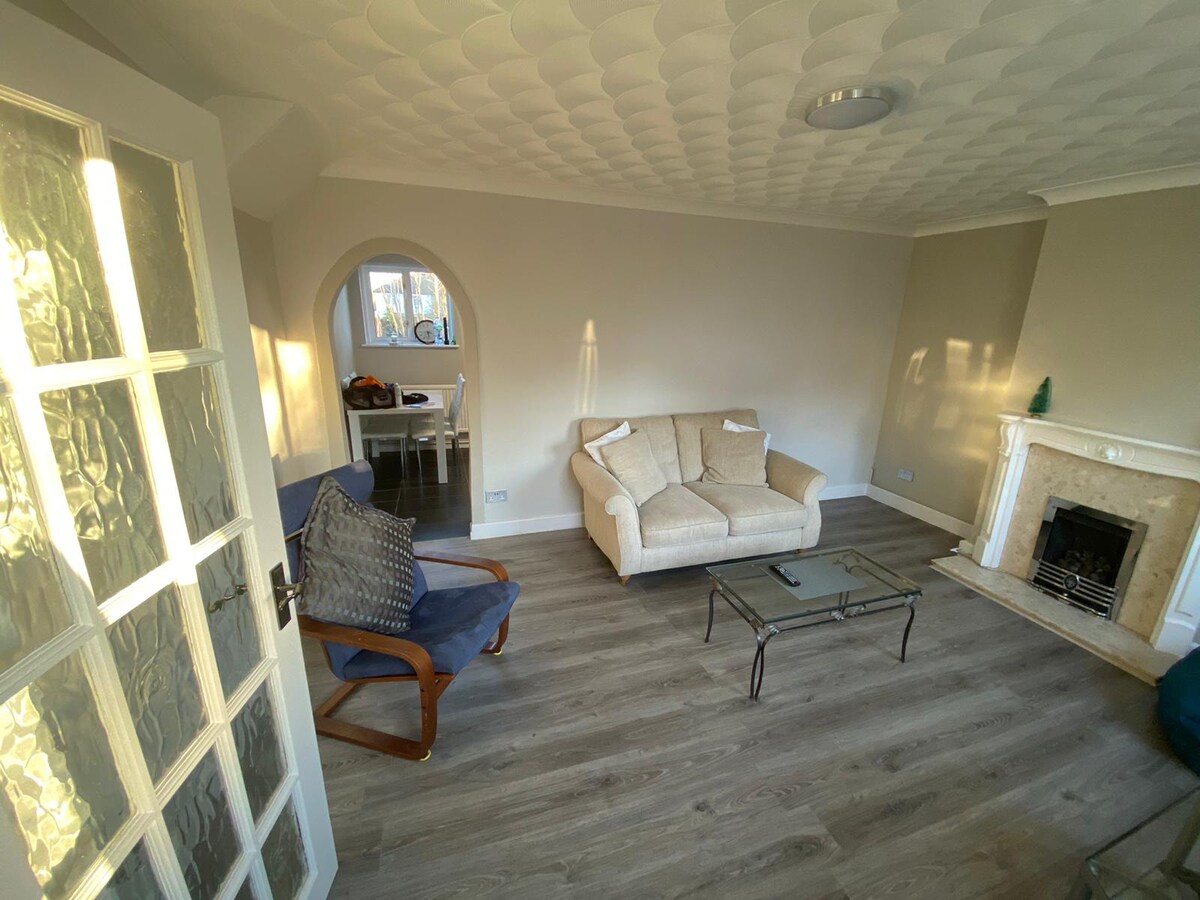 The spacious lounge is fitted with a light-colored sofa and a glass coffee table, creating a comfortable seating area. Natural light enters through a nearby window, highlighting the modern décor. A cozy armchair is placed beside the fireplace, enhancing the room's relaxing ambiance.