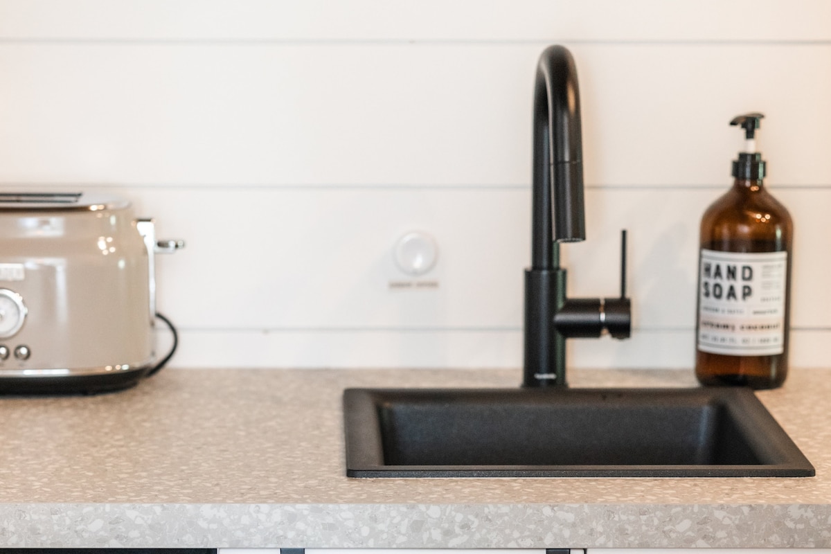 A modern kitchen countertop is visible, featuring a sleek black sink and a contemporary faucet. Nearby, a toaster sits in a soft hue and a glass soap dispenser labeled 'HAND SOAP' stands against the light-colored wall.