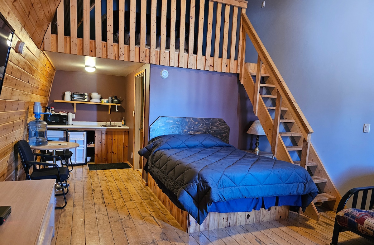 A spacious room features a double-level layout with wooden walls and a loft area visible above. A large bed with a dark quilt is positioned against the back wall. A kitchenette with modern appliances and a small dining area is nearby, with stairs leading to the loft.