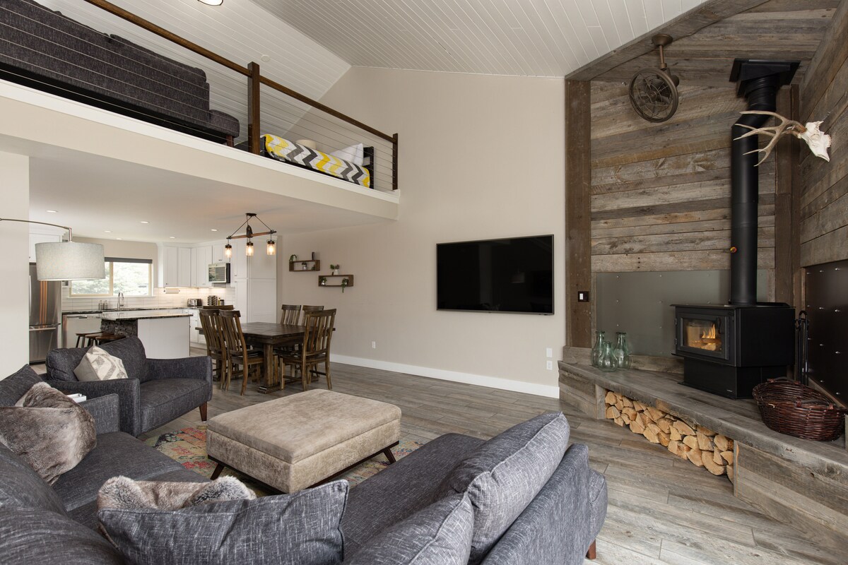 A spacious living area features comfortable seating with a large sectional sofa. A wood stove is positioned against a rustic wall, complemented by a flat-screen TV. A loft area is visible above, and a dining table is set nearby, creating an inviting gathering space.