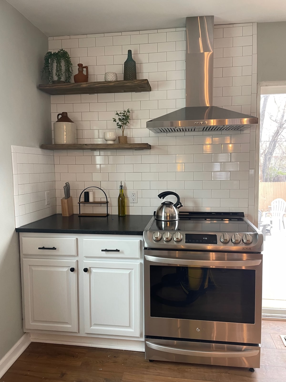 The kitchen features a sleek stainless steel stove and hood, complemented by white cabinets. Open shelving displays decorative items and a plant. A neutral color palette contributes to a modern and inviting space.