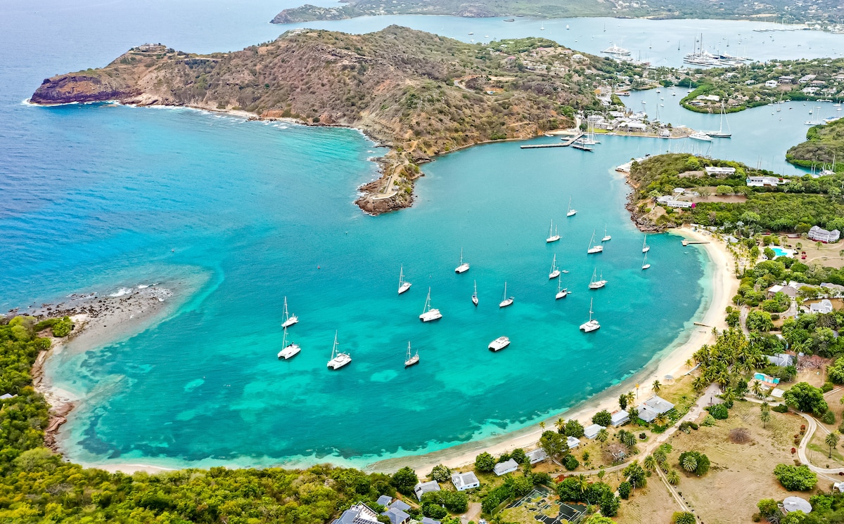 An aerial view captures a vibrant bay surrounded by lush greenery and rolling hills. Sailboats are anchored in the calm turquoise waters, with sandy beaches lining the shore. The scene highlights the serene coastal landscape and the variety of boats dotting the harbor.