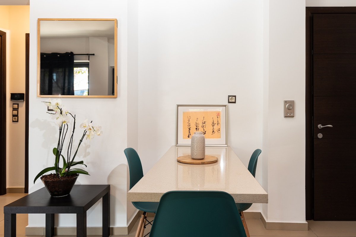 A minimalist dining area is displayed, featuring a light-colored table with modern design. A decorative vase holds fresh flowers, while a framed artwork adds a subtle touch to the wall. A mirror is mounted nearby, enhancing the sense of space.